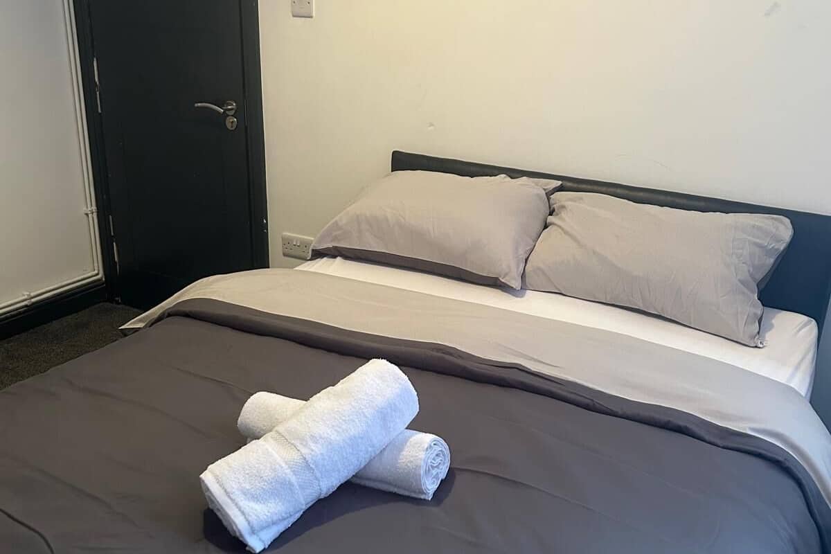 Un lit bien fait, avec une literie grise et deux serviettes blanches roulées, une porte sombre et un mur blanc uni.