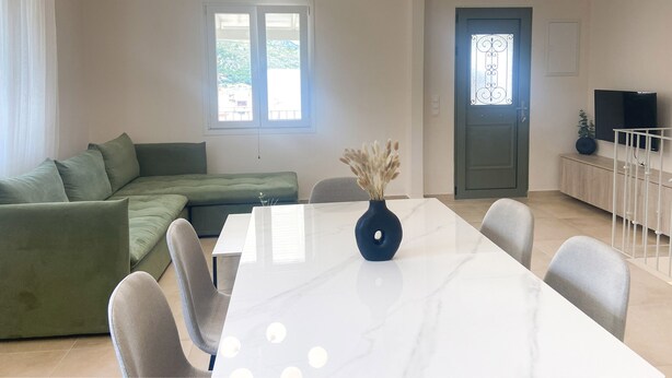 A modern living room with a green sectional sofa, a white dining table with chairs, a vase with dried pampas grass, and a TV on a wooden cabinet. - Nonna's Hill House (Corfu)