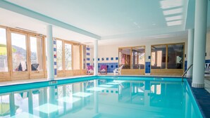 Una piscina interior con paredes de vidrio transparente, terraza de madera y sillas de descanso.