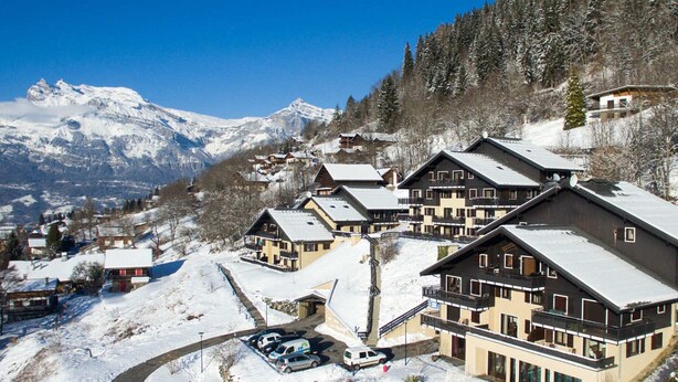Un pueblo en la montaña cubierto de nieve, con edificios de estilo chalete, un área de estacionamiento y una vista despejada de las montañas circundantes.