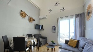 Una sala de estar con un sofá gris, una mesa de comedor con sillas, un televisor y un balcón con cortinas.