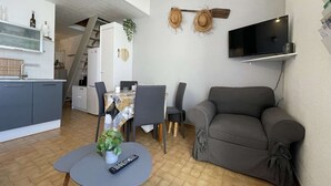 Un espacio habitable compacto con zona de comedor, sofá, televisor montado en la pared y cocina con refrigerador y pileta.