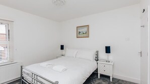 Un dormitorio blanco con una cama, mesitas de noche, un radiador, un cuadro en la pared y una ventana con persianas.