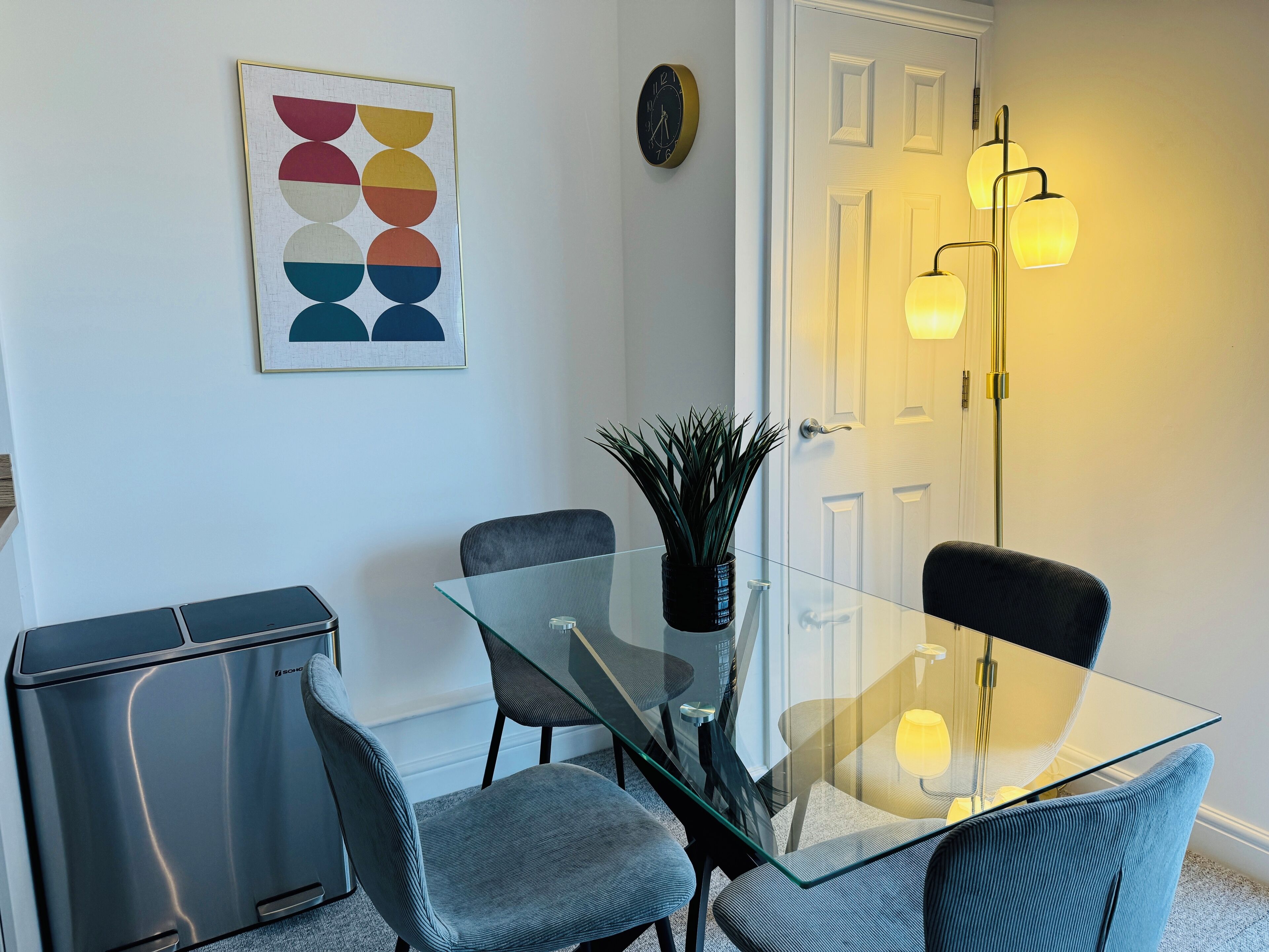 Un comedor moderno con una mesa de vidrio, dos sillas grises tapizadas, una planta en maceta y un reloj montado en la pared.