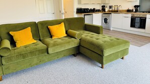 A green sectional sofa with yellow cushions in a modern living room with a kitchen in the background. - Spacious One Bed Apt With Scenic Views (Ventnor)