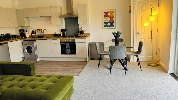A modern kitchen with white cabinets, a stainless steel range, and a built-in oven. There is a dining area with a glass table and chairs, and a green upholstered sofa. - Spacious One Bed Apt With Scenic Views (Ventnor)