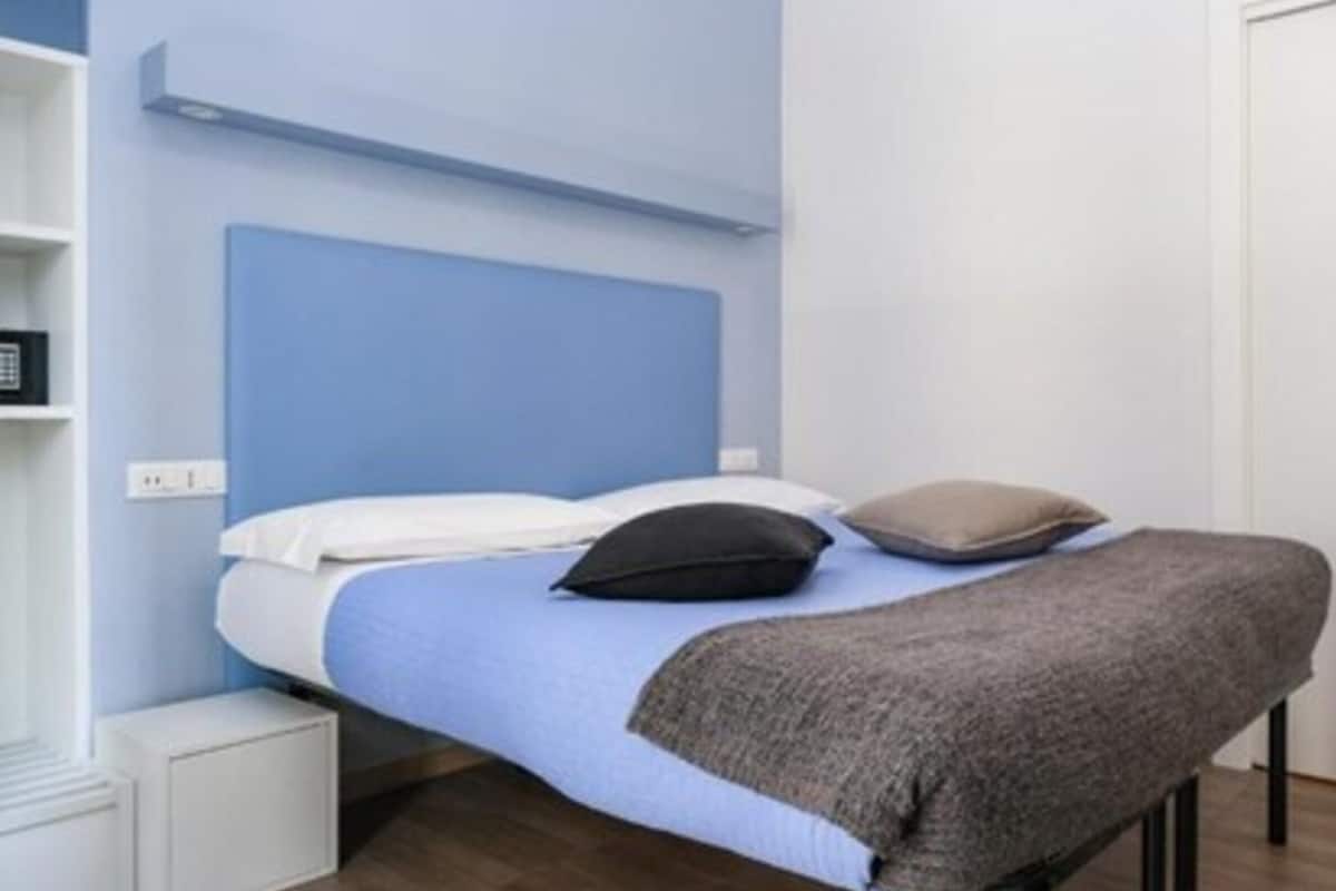 Een eenpersoonsbed met een blauw hoofdbord, witte beddengoed en twee kussens. Links van het bed staat een witte nachtkastje.