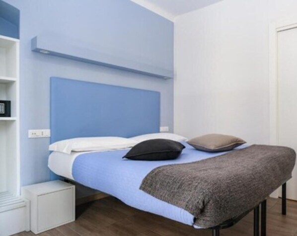Een eenpersoonsbed met een blauw hoofdbord, witte beddengoed en twee kussens. Links van het bed staat een witte nachtkastje.
