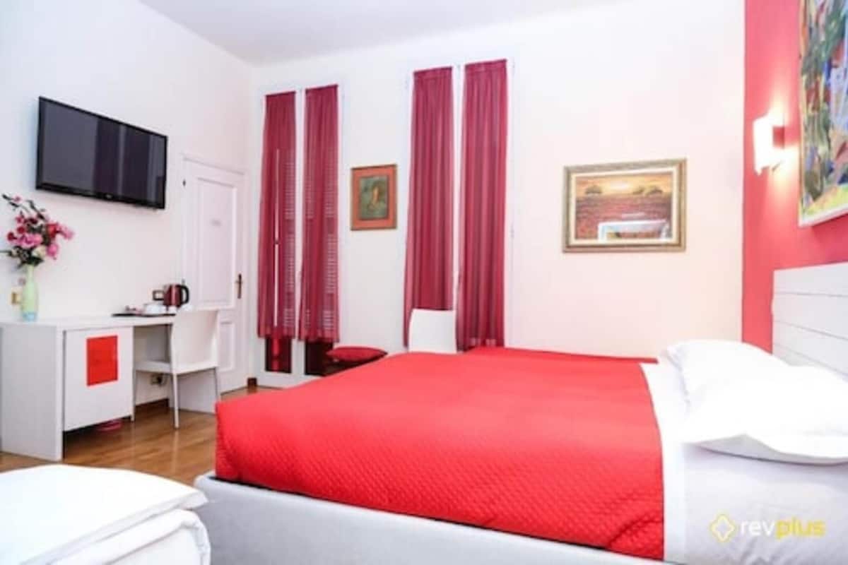 Een hotelkamer met een bed, een bureau, een stoel, een televisie en twee ramen met rode gordijnen.