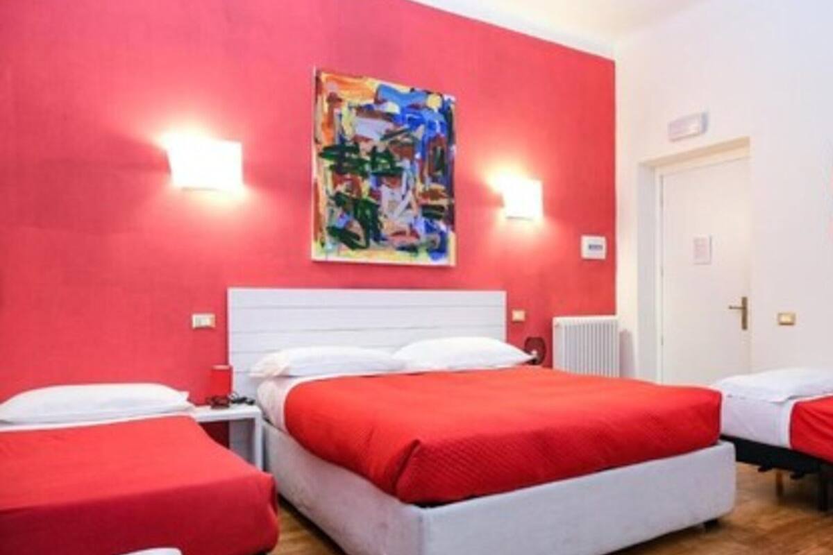 Hotelkamer met een rode wand, een wit hoofdbord en een schilderij boven het bed. Er staan twee bedden met rode beddengoed en witte kussens.