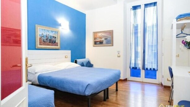 A bedroom with two beds, a blue wall, a door with curtains, and a small table with a chair. - Lia Rooms (La Spezia)
