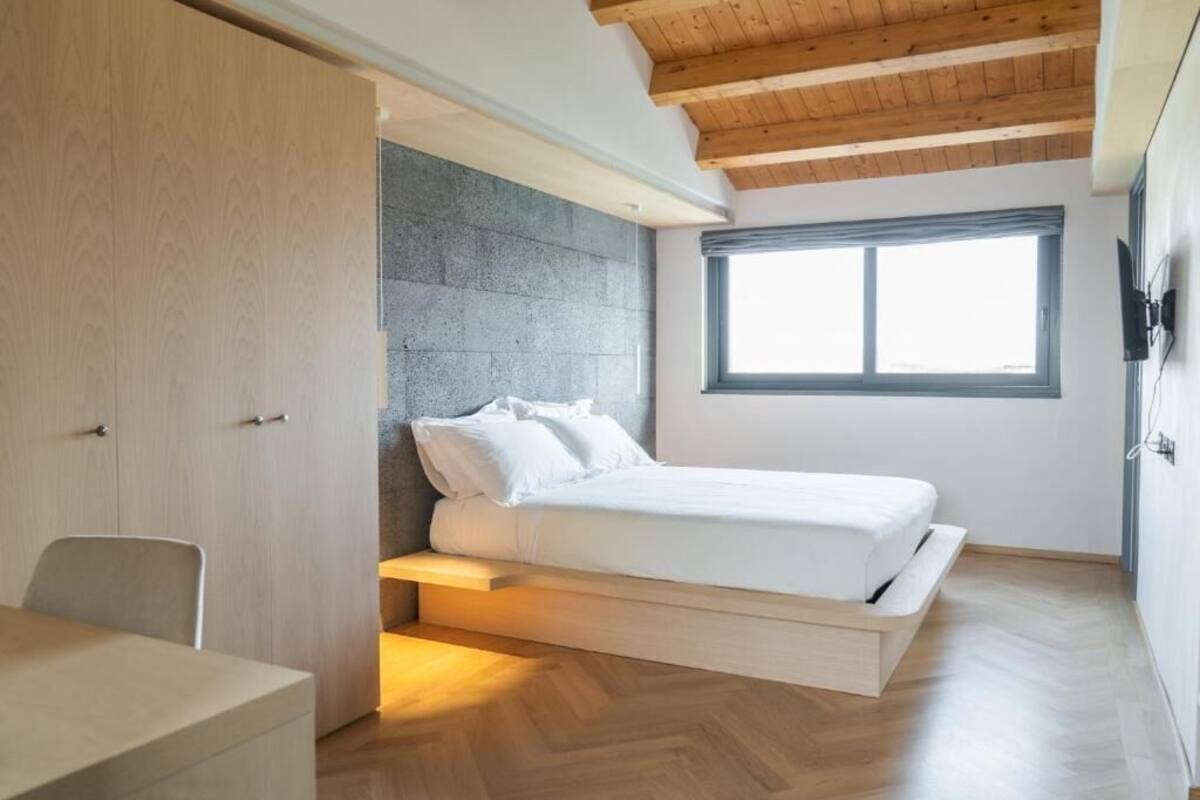 A bedroom with a bed, a desk, a chair, a window, and a wooden ceiling.