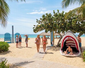Une scène de plage avec un espace de jeux en forme de requin, des chaises de plage et une vue dégagée sur l’océan.