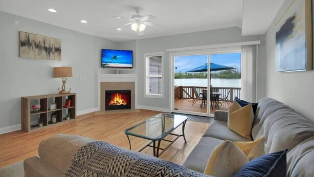 A living room with a fireplace, a television, and a balcony with a view of a mountain and water. - Lakefront Beautiful Townhome w/ Stunning Views! (North Charleston)