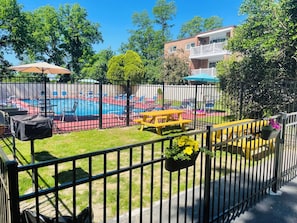 Ein von einer Hecke umgebener Poolbereich mit Liegestühlen und einem Tisch, umgeben von Grünpflanzen und einem Gebäude im Hintergrund.