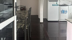 Um corredor estreito com um espaço de jantar de um lado, com uma mesa e cadeiras. Uma geladeira branca é visível ao fundo, e o chão é de madeira escura.