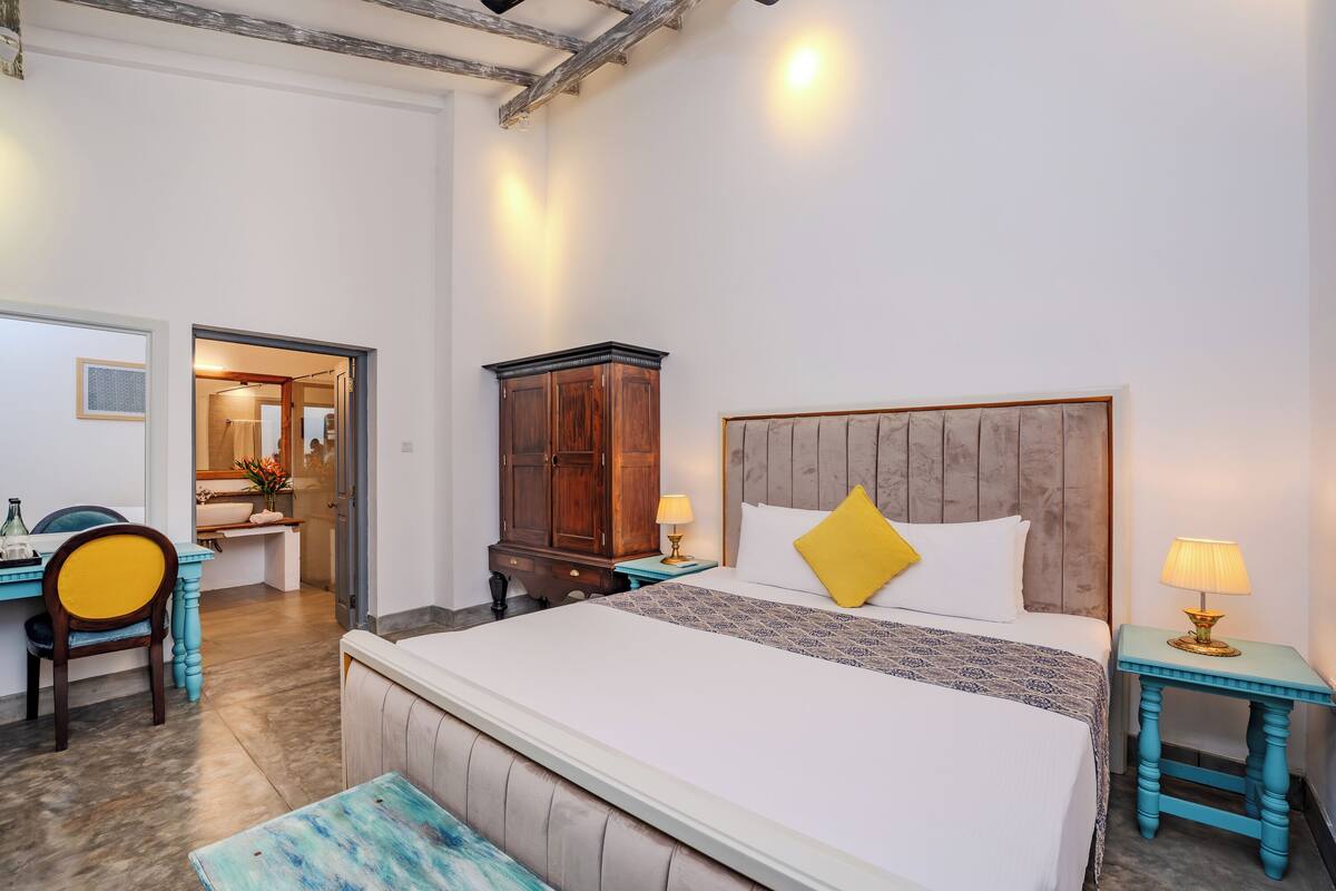 A hotel room with a large bed, a desk with a chair, a wardrobe, and a bathroom visible through an open door.