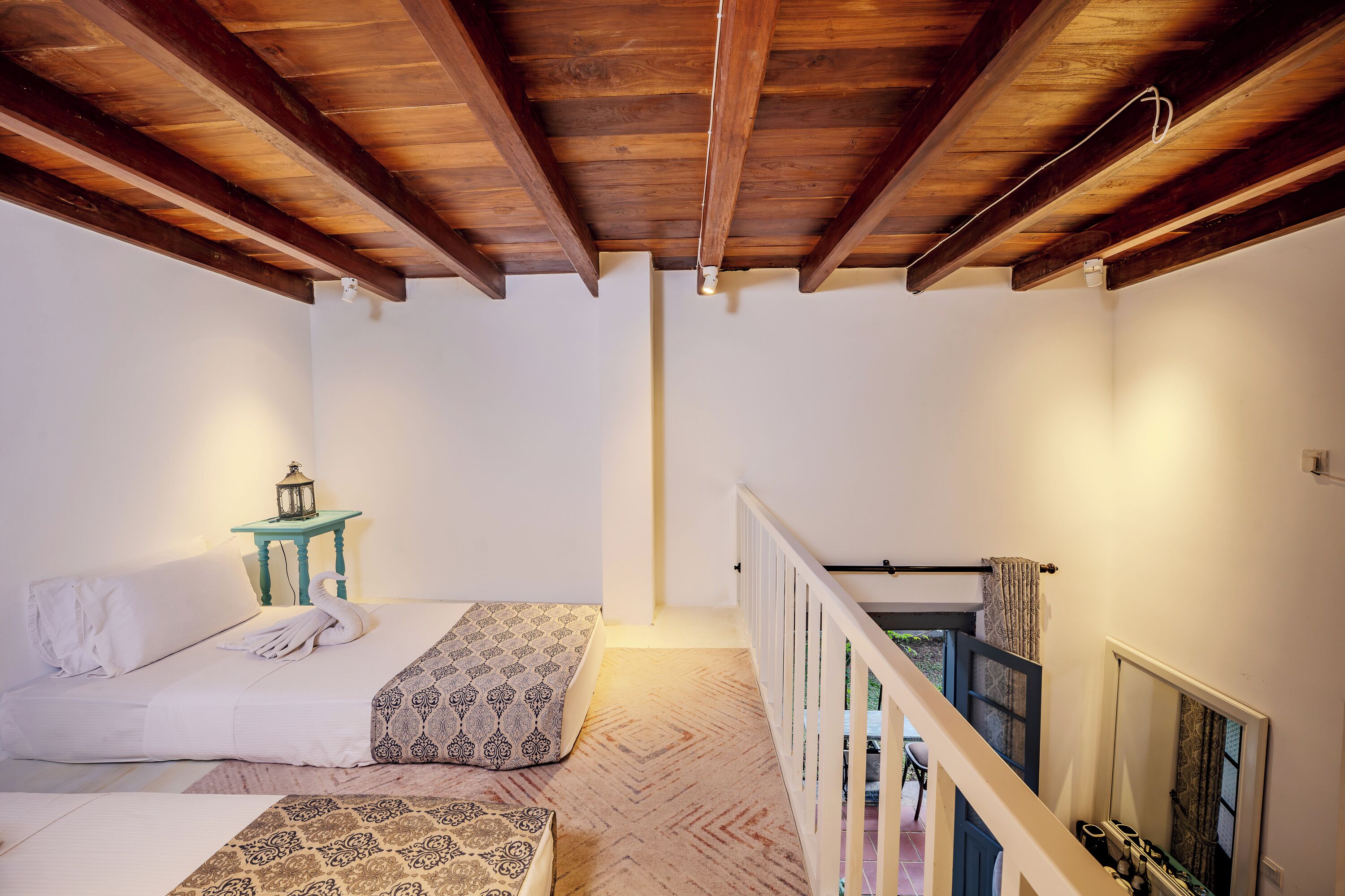 A bedroom with two beds, a wooden ceiling, a small table with a lantern, and a view of a balcony.