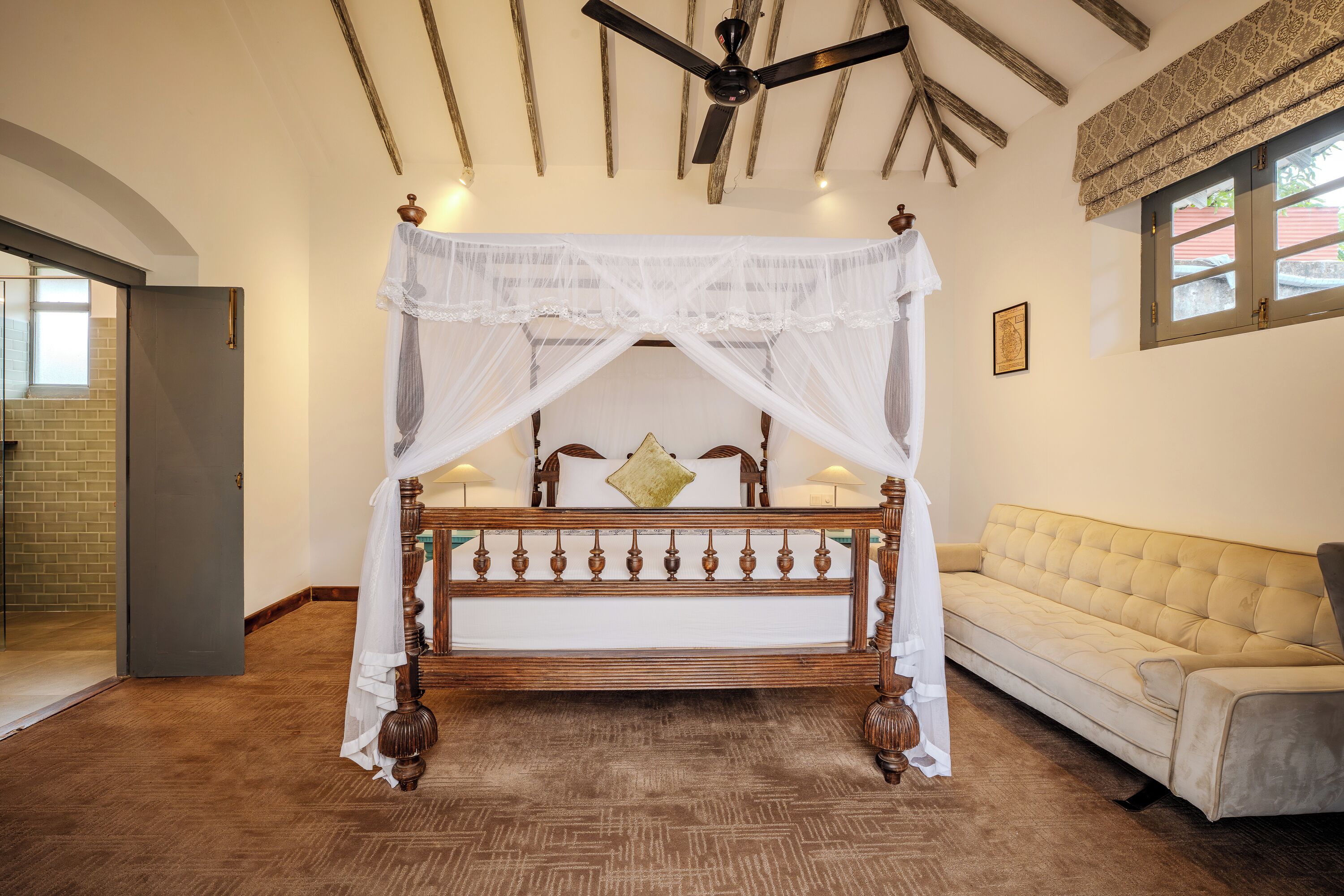 A bedroom with a four-poster bed, a ceiling fan, a beige sofa, and a patterned window valance.