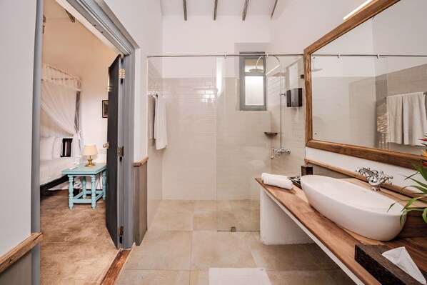 A modern bathroom with a large mirror, a wooden vanity, a shower, and a small bedside table with a lamp.