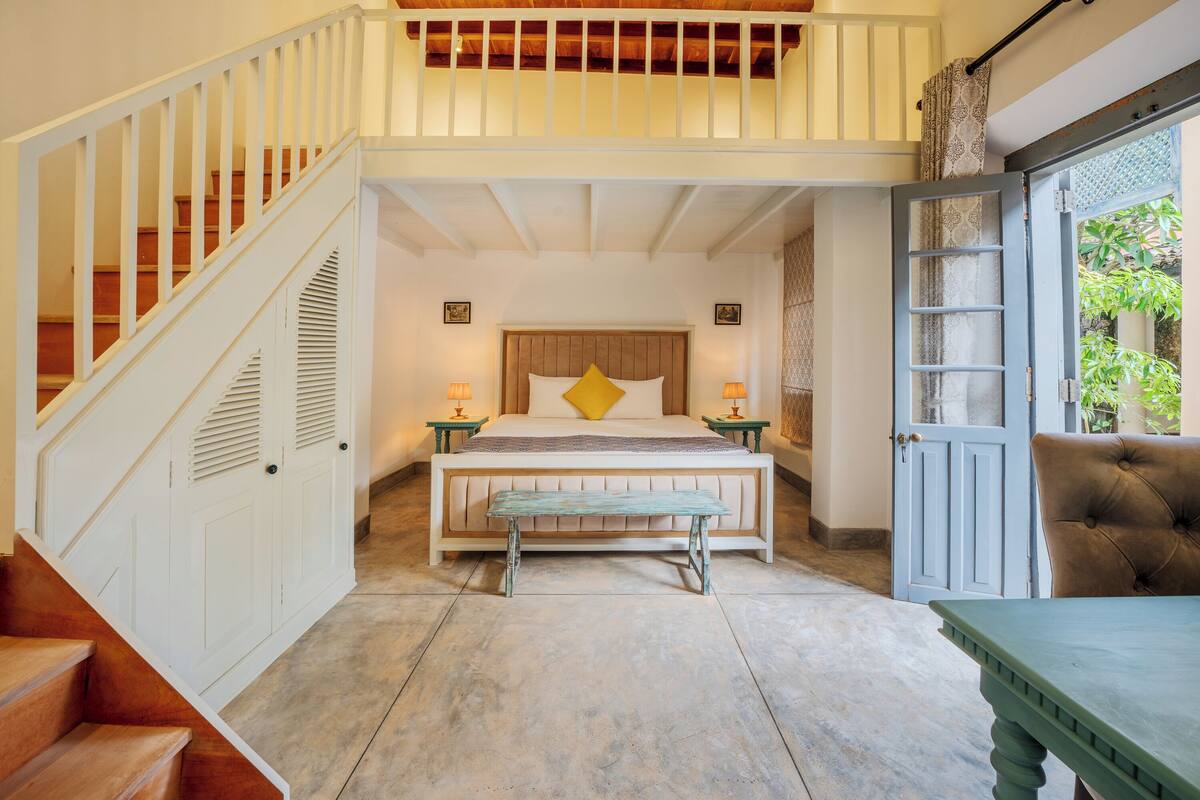 A bedroom with a wooden staircase, a bed with a wooden headboard, a bench, and a green table.