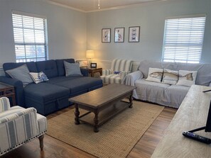 Un salon avec un canapé bleu, un fauteuil à rayures et une table basse en bois. Il y a deux fenêtres avec des stores, une lampe et des œuvres d’art encadrées accrochées au mur.