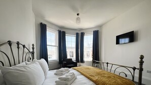 Una cama bien hecha con sábanas blancas y una manta amarilla, una silla negra, un televisor de pantalla plana en la pared y una ventana con cortinas.