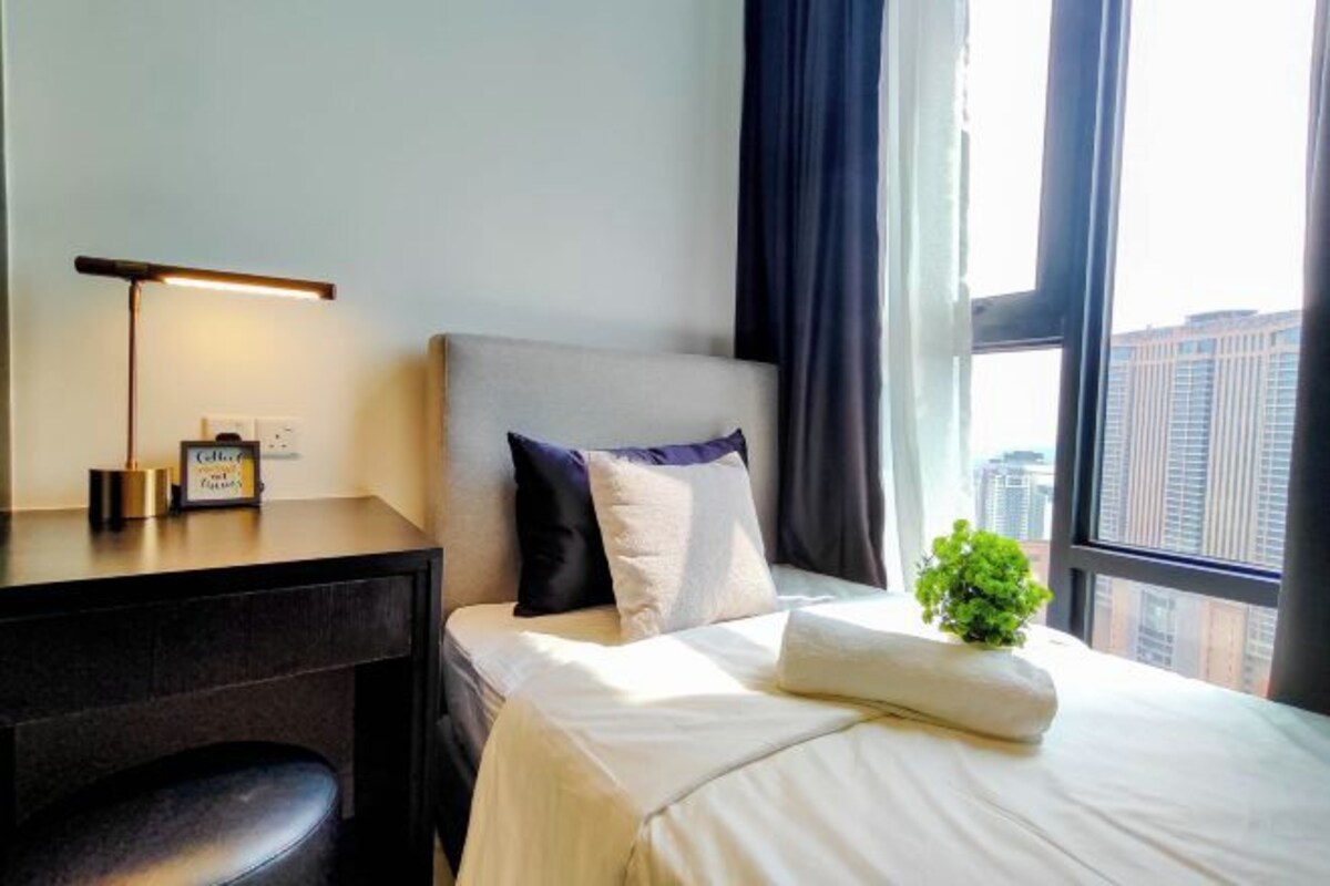 A hotel room with a bed, a desk with a lamp, a small plant, and a window with a city view.