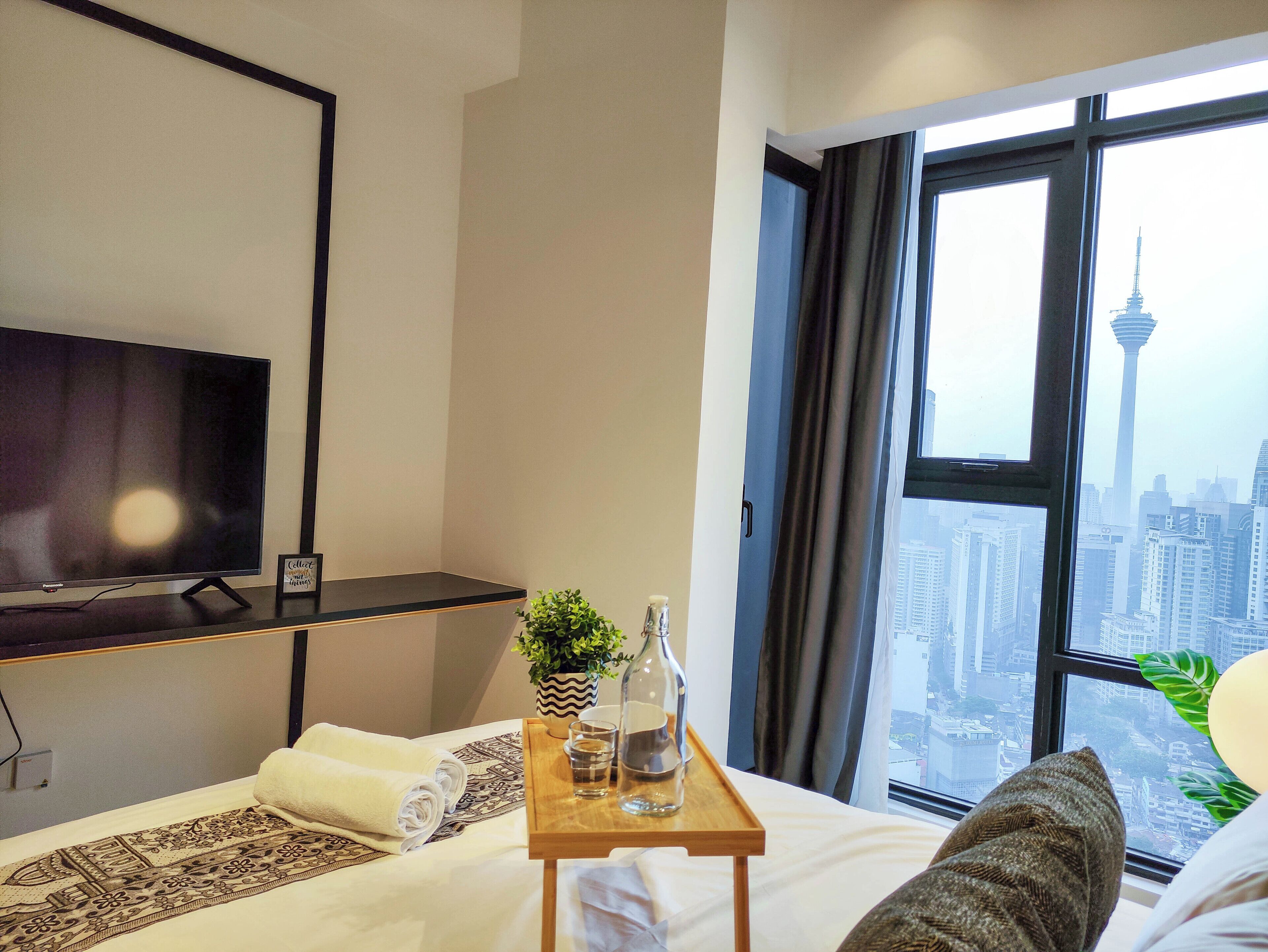 A hotel room with a bed, a TV, a window with a city view, and a tray with a bottle and glasses.