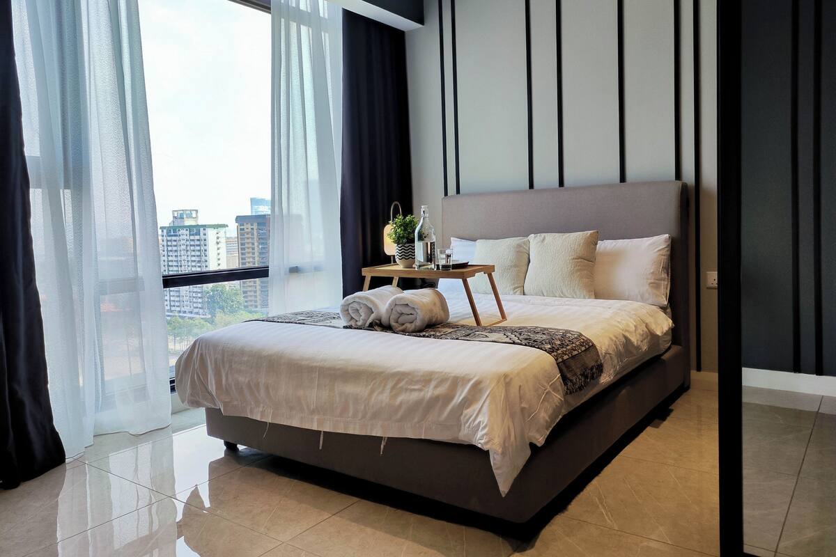 Un dormitorio moderno con una cama grande, una mesita de noche con lámpara y vistas a la ciudad a través de amplias ventanas.