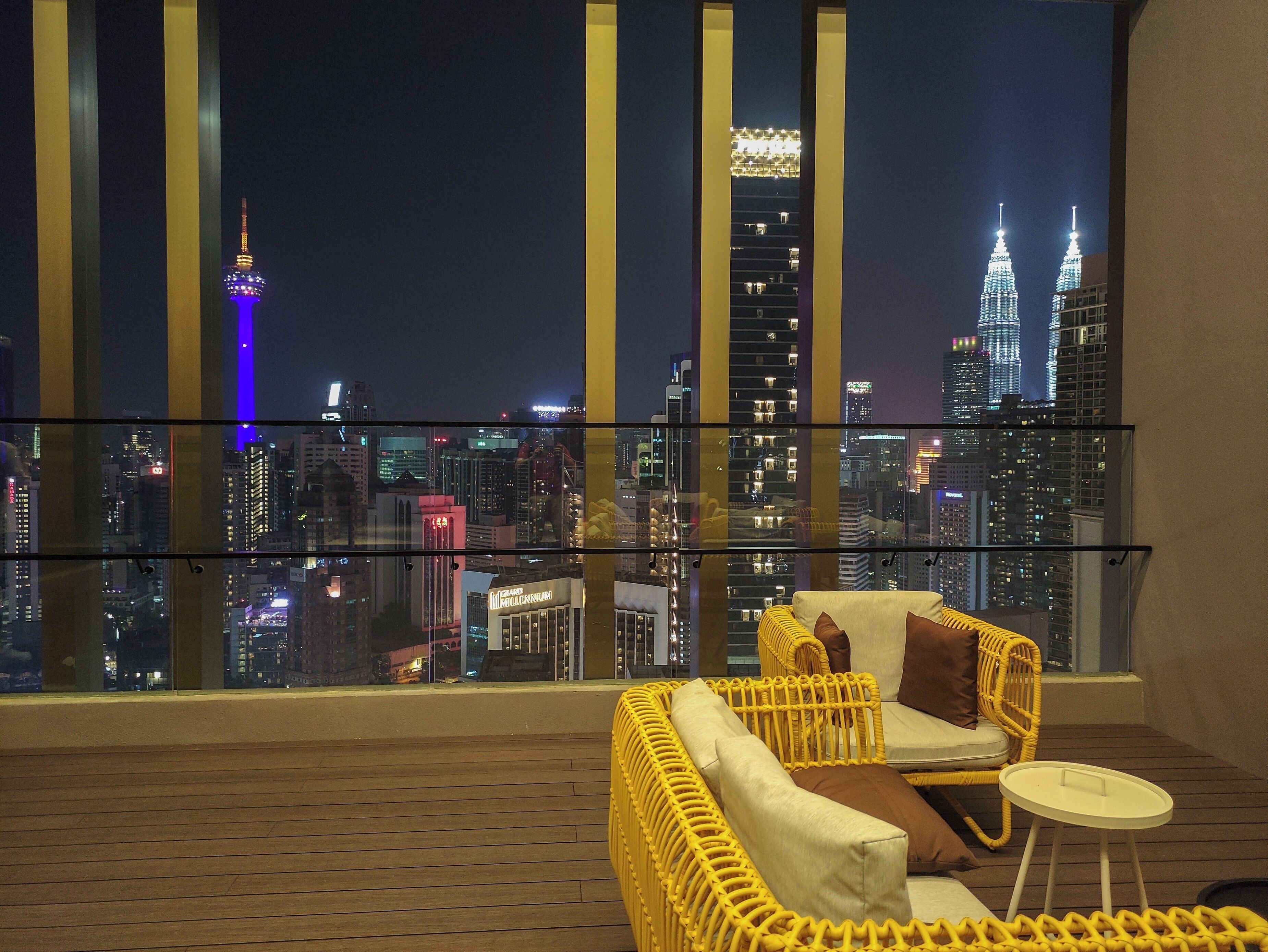 A balcony with a yellow wicker sofa, a small round table, and a city view at night.