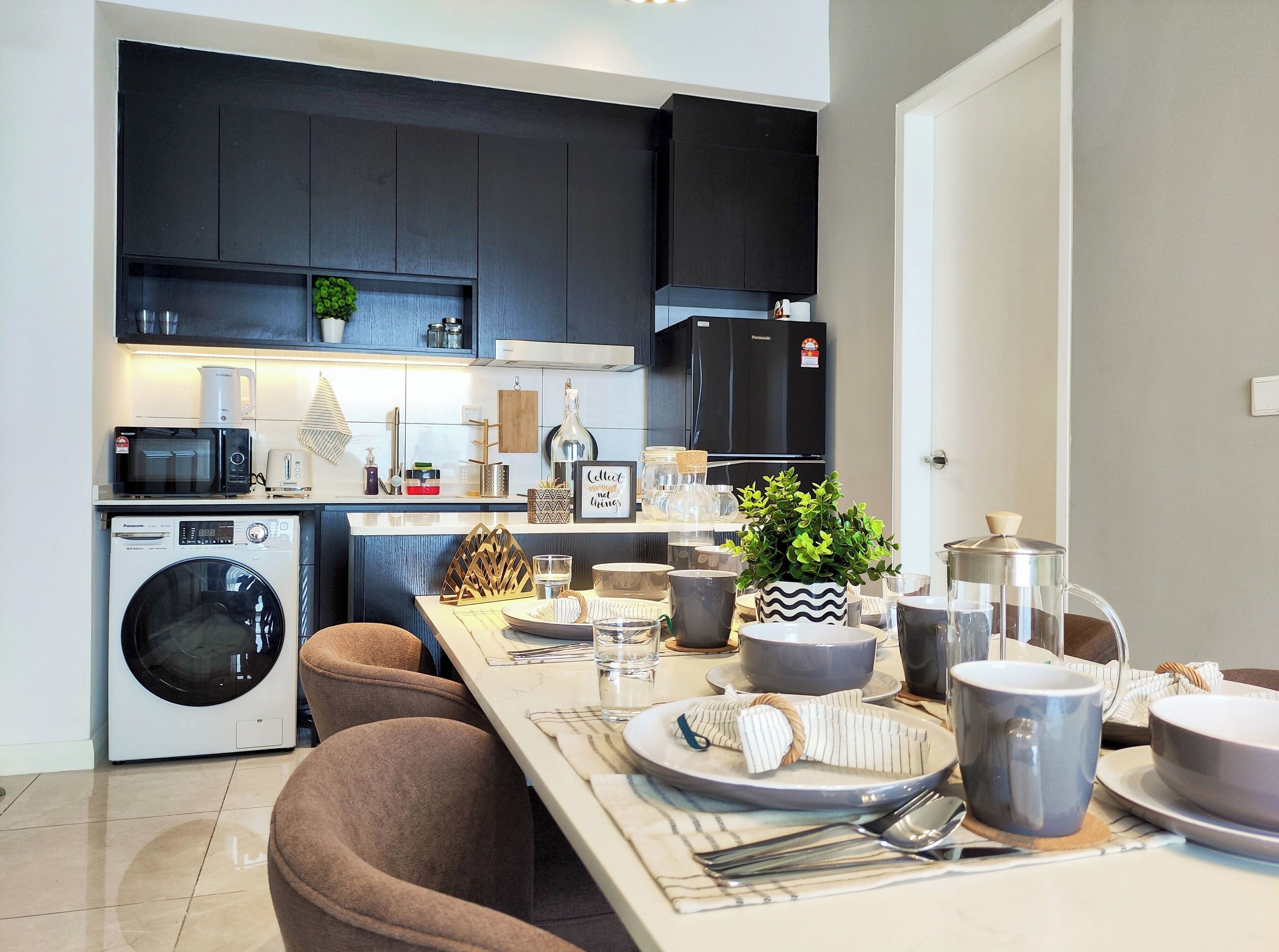 A modern kitchen with dark cabinets, a washing machine, a dining area with a table set for two, and a refrigerator.