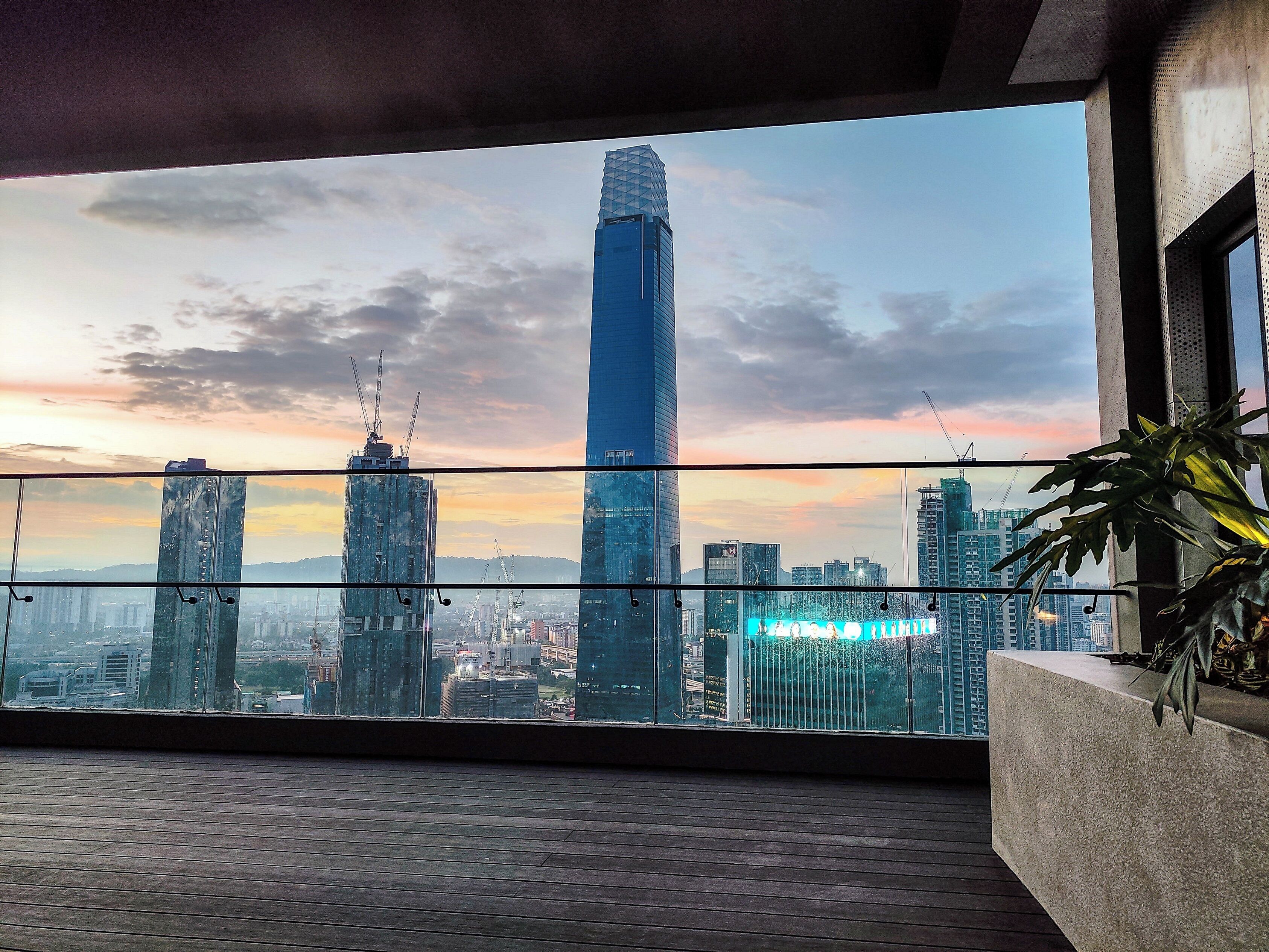 Un balcón con vistas a un paisaje urbano, que incluye un edificio alto de diseño único y varios otros rascacielos.