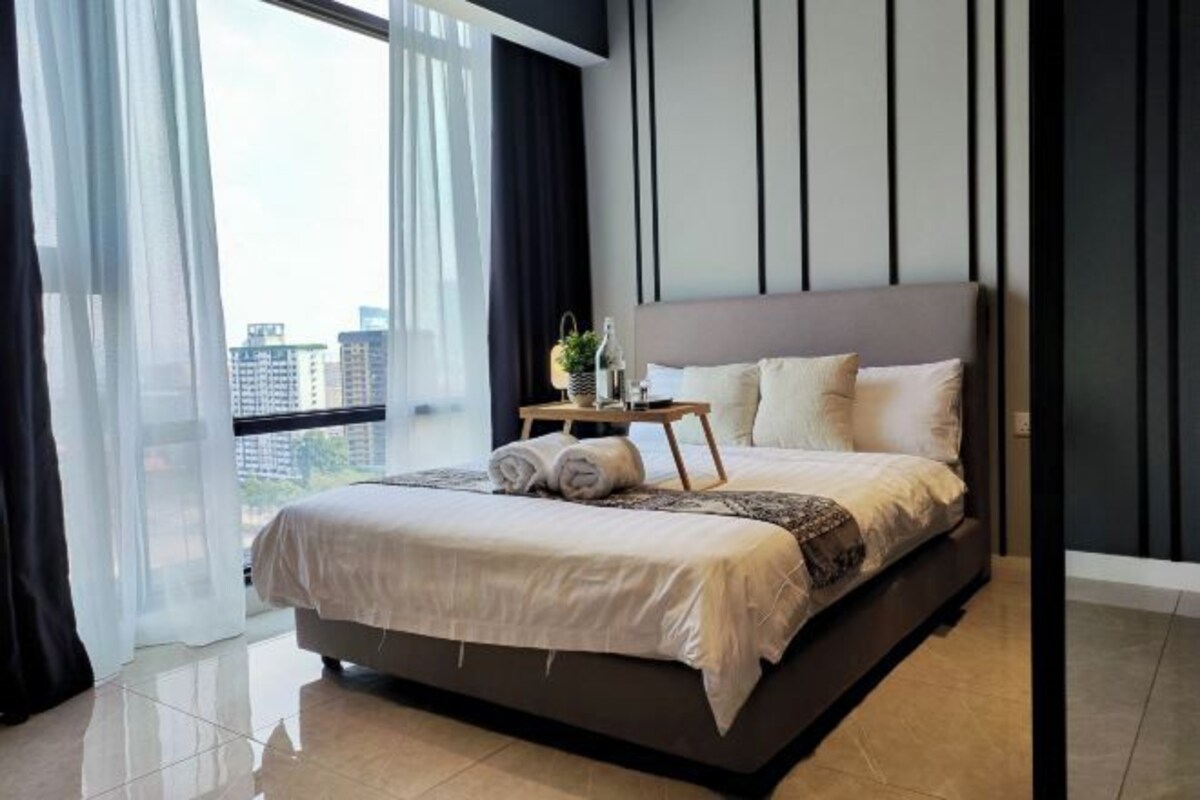 A modern bedroom with a large bed, a bedside table with a lamp, and a view of the city through sheer curtains.