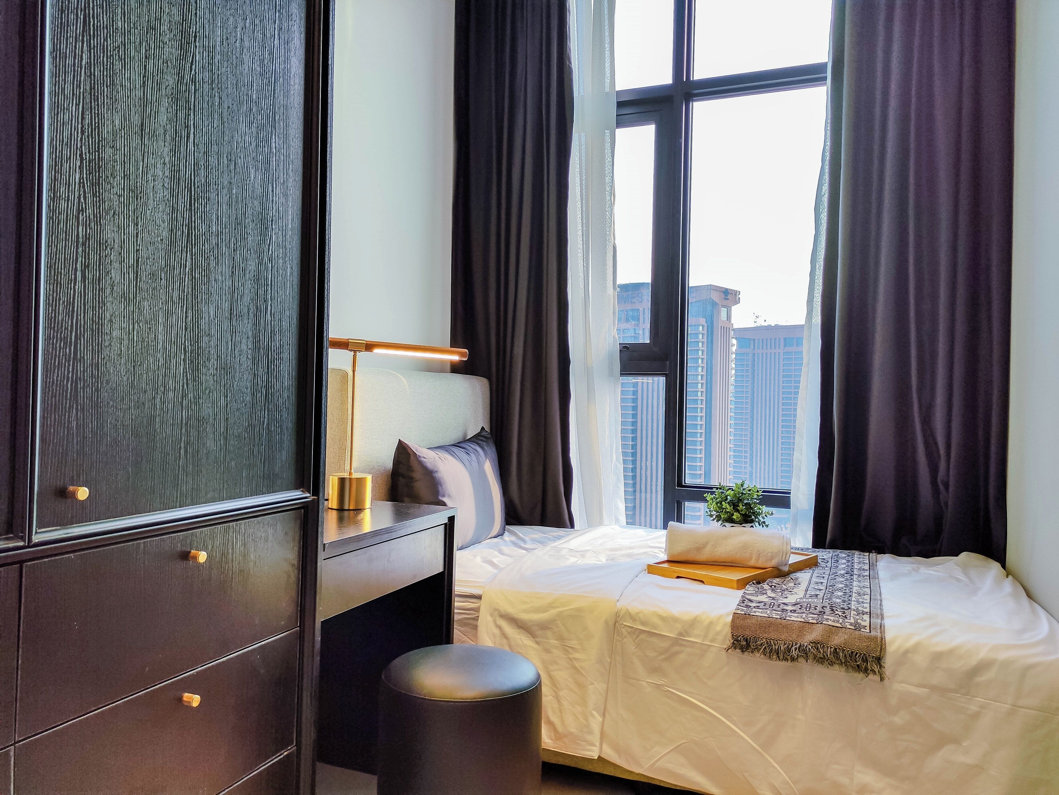 A modern hotel room with a large window, a bed, a desk, and a dark wooden wardrobe.