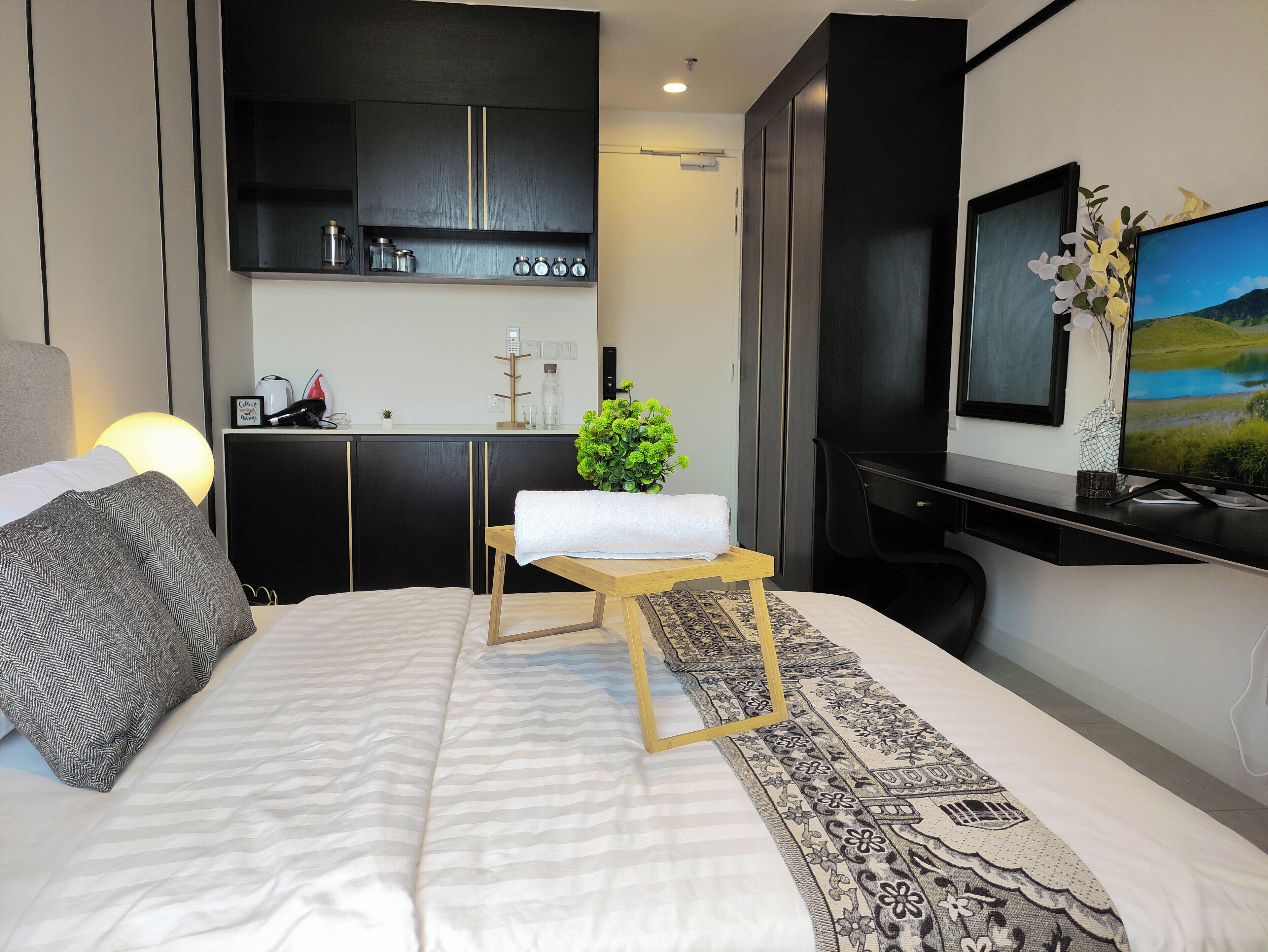 Una habitación de hotel moderna con una cama grande, una pequeña mesa de madera con una toalla, un escritorio con una computadora, un televisor y una zona de cocina al fondo.