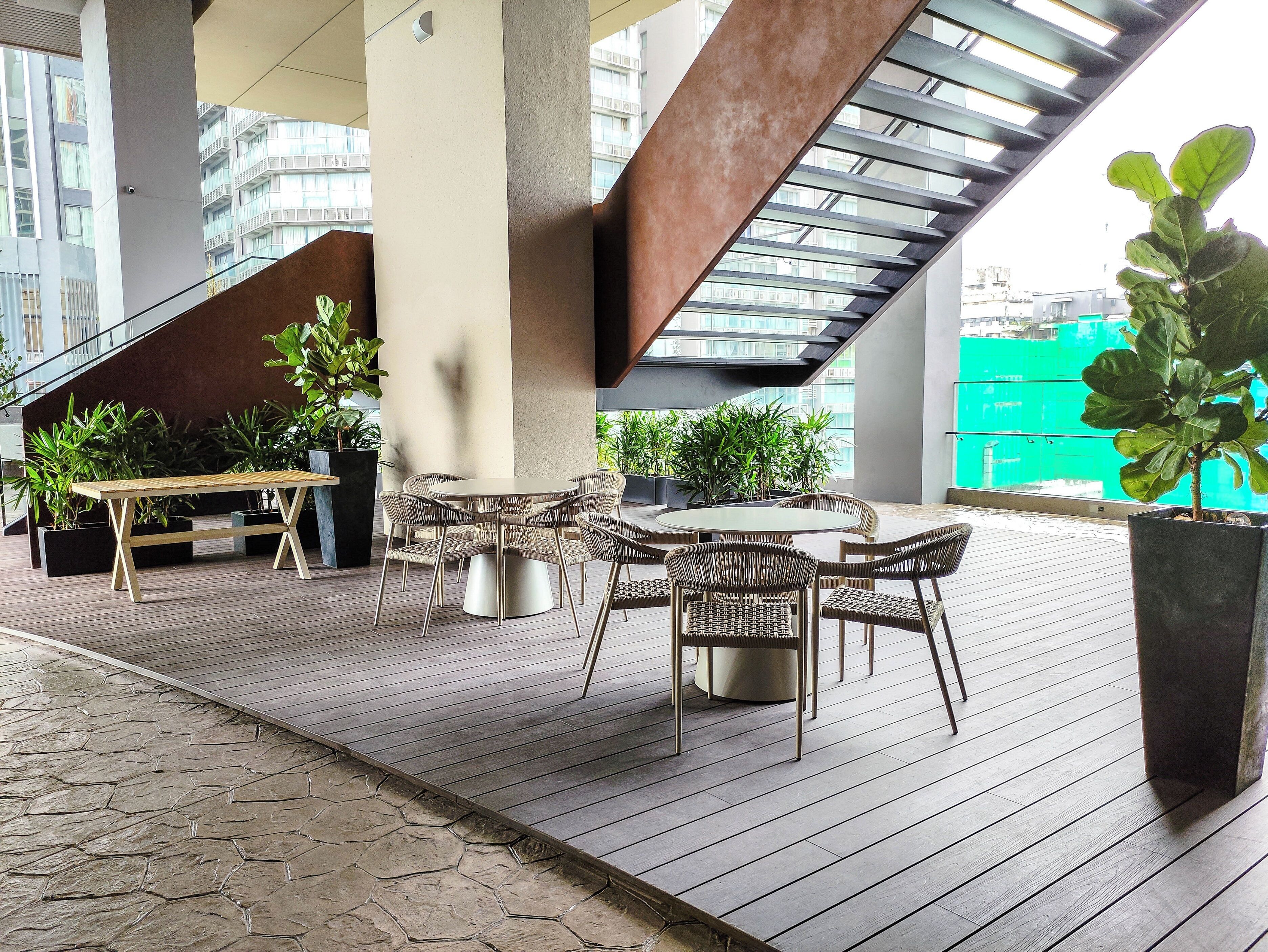 Un área de estar al aire libre con una mesa redonda y sillas de mimbre, rodeada de plantas en macetas, bajo una estructura arquitectónica moderna con techo de vidrio.