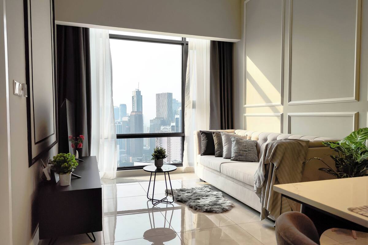 Una sala de estar moderna con un sofá, una mesita redonda y vistas a un paisaje urbano a través de amplias ventanas.