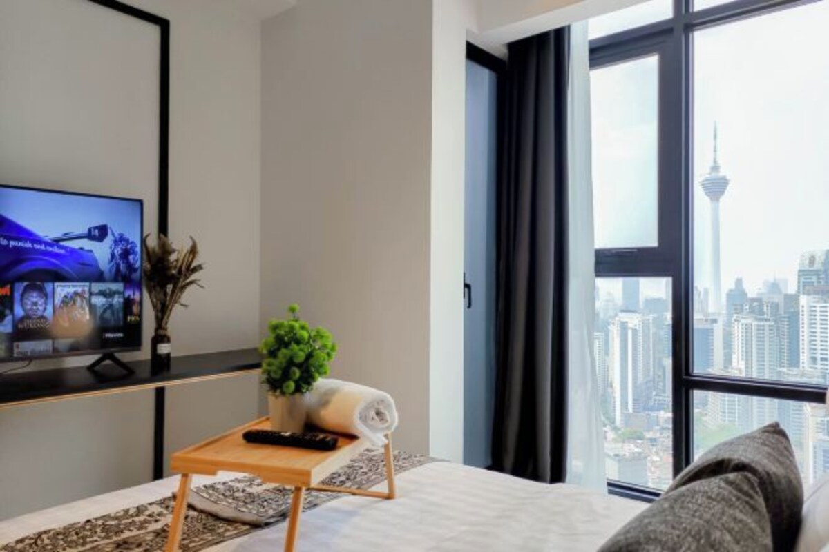 A modern hotel room with a large window offering a city view, a bed with a white headboard, a small wooden table with a remote control, a rolled towel, and a potted plant.