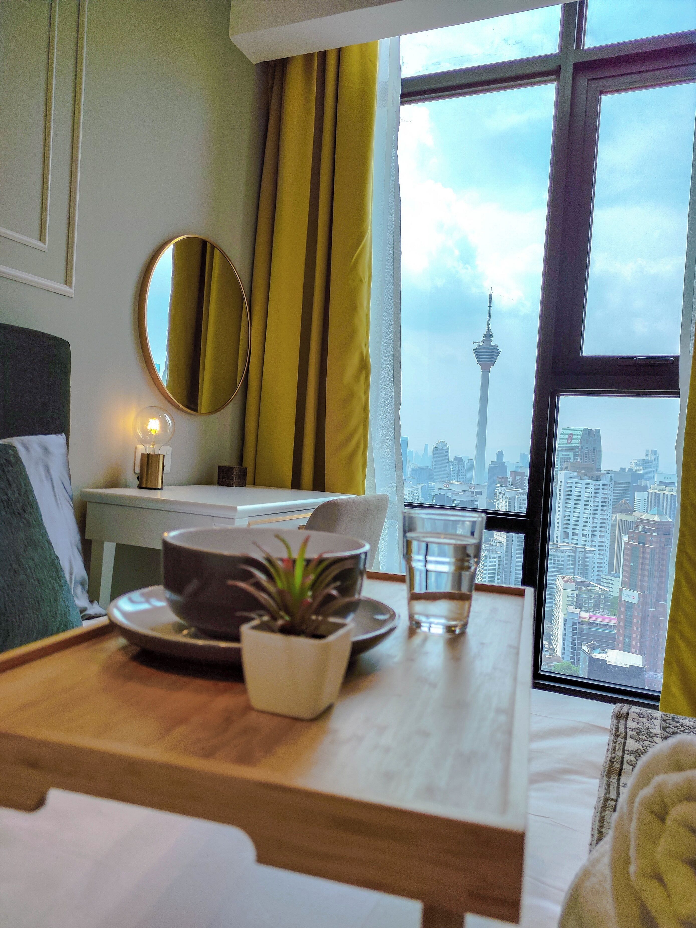 A hotel room with a bed, a bedside table with a lamp, a window with a city view, and a view of a tower.