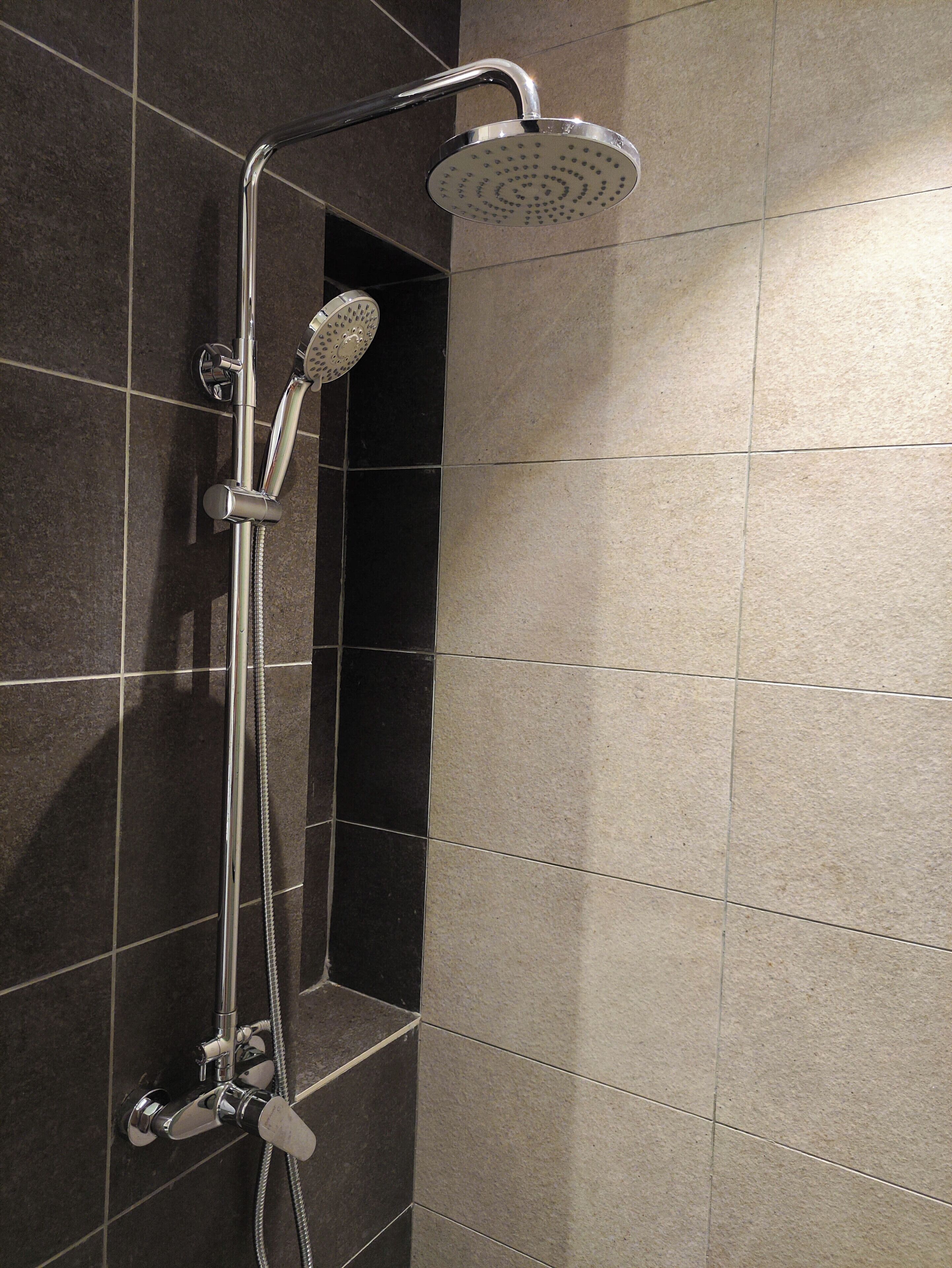 A shower with a handheld showerhead and a fixed overhead showerhead, mounted on a wall with dark and light tiles.