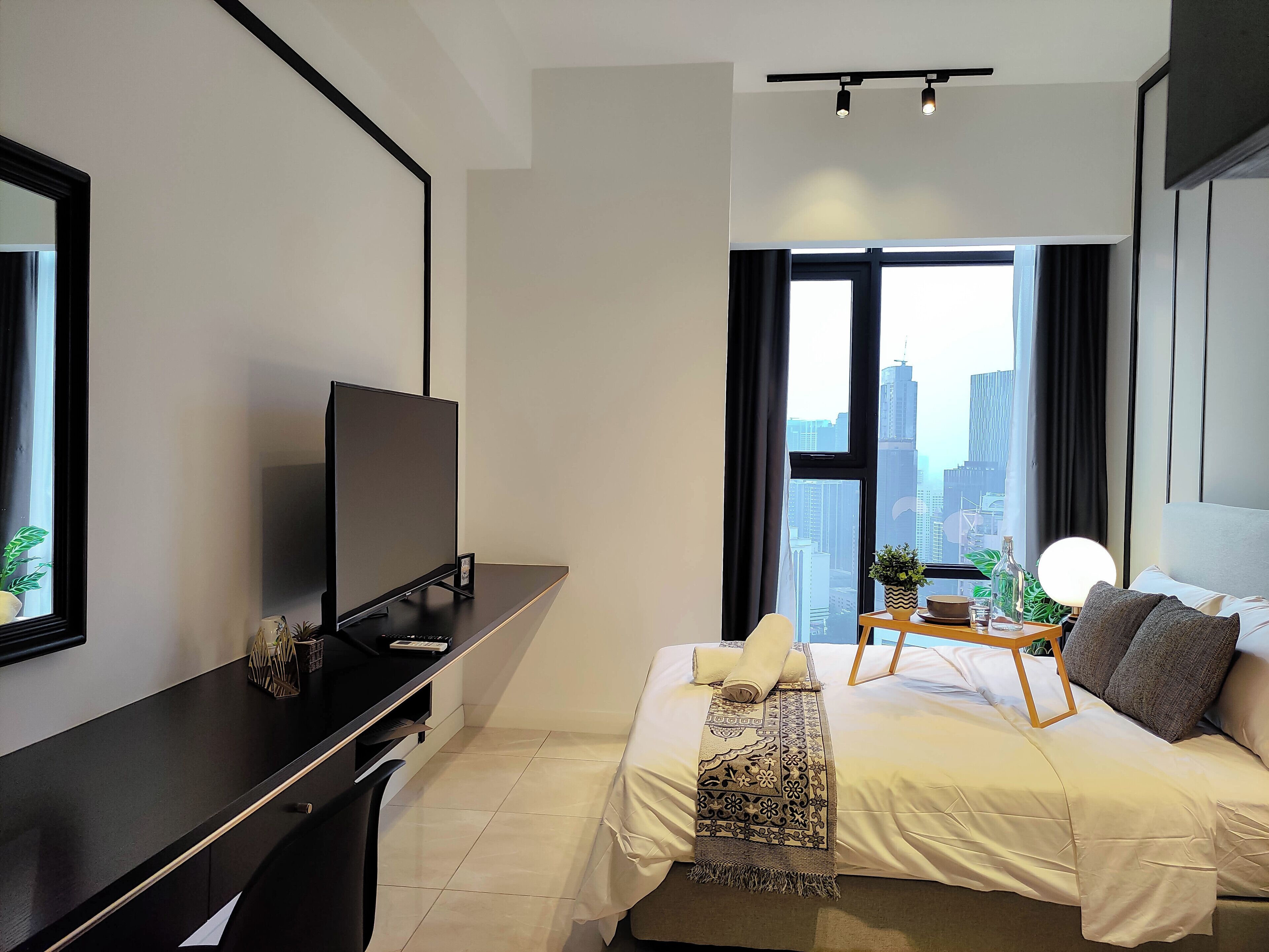 A modern hotel room with a large bed, a desk with a flat-screen TV, a bedside table with a lamp, and a view of the city skyline through large windows.