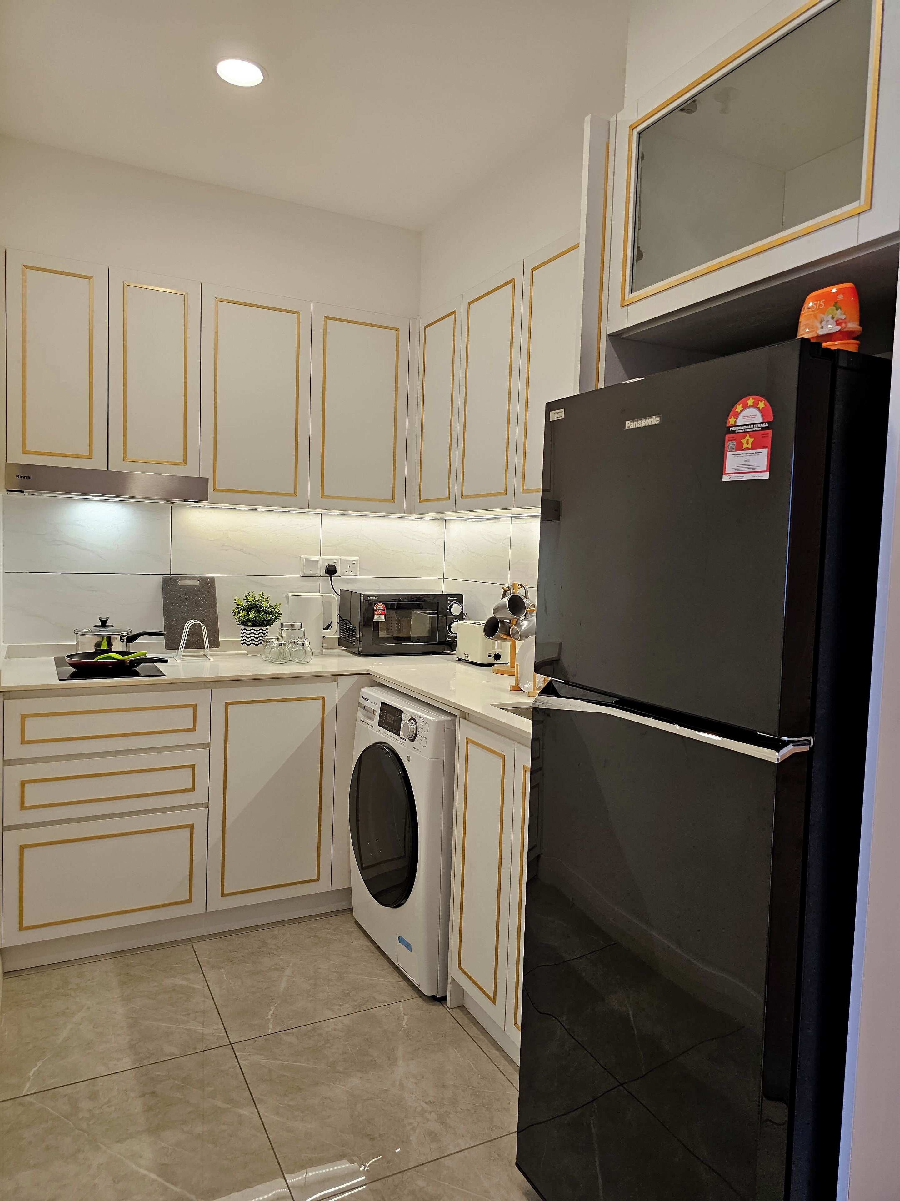 Una cocina compacta con armarios blancos, un refrigerador negro, un microondas, una lavadora y un lavamanos.