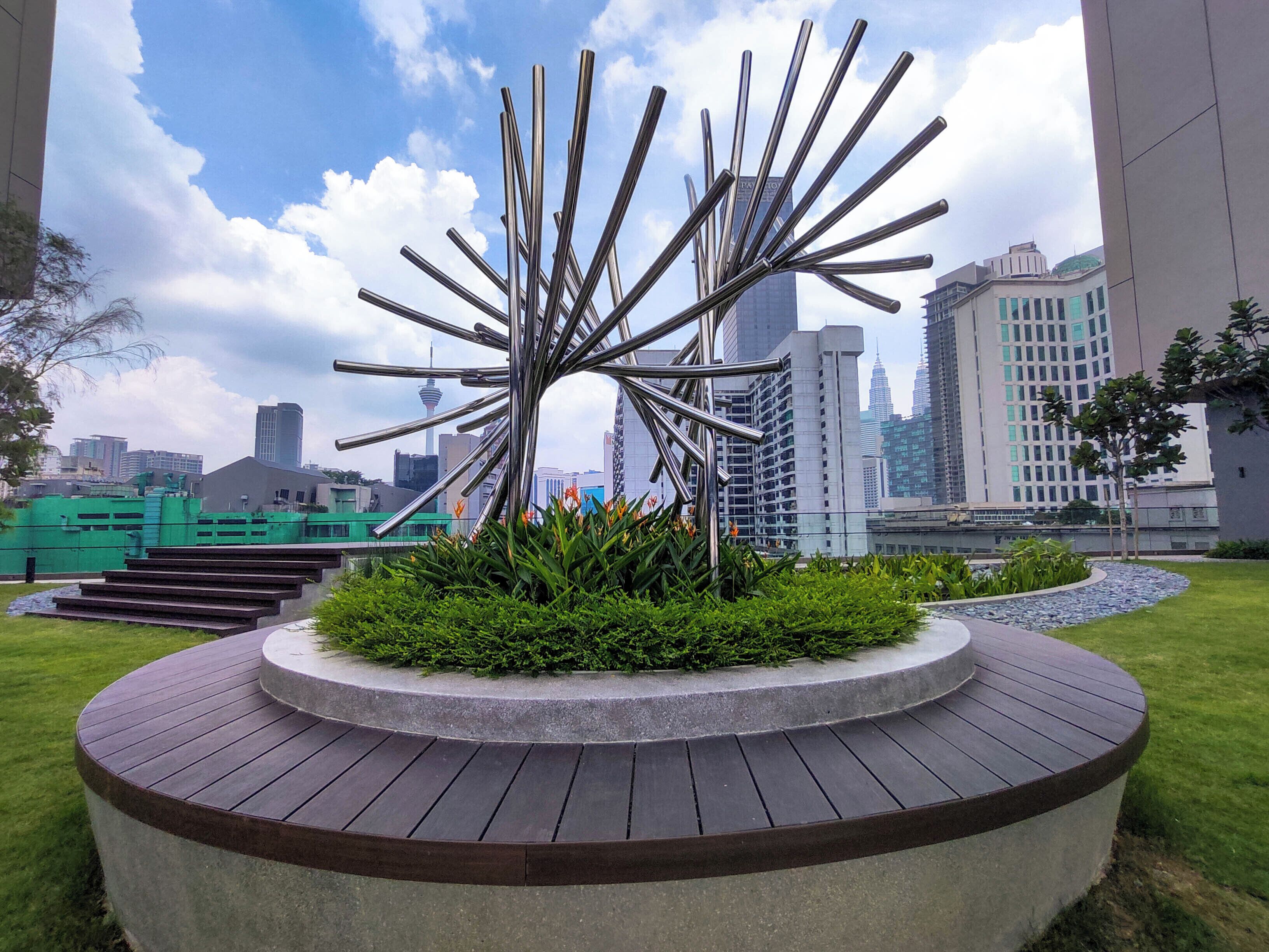 A modern sculpture in a circular concrete planter surrounded by a wooden deck, with a cityscape in the background.