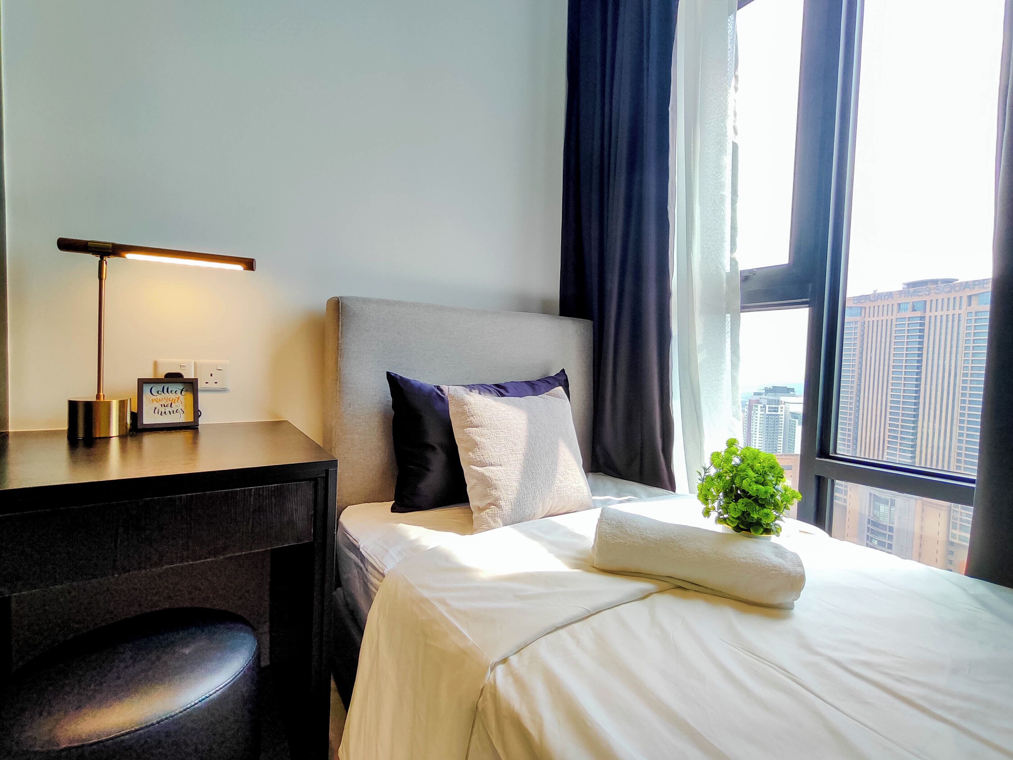 A hotel room with a bed, a desk with a lamp, a small plant, and a view of buildings through the window.