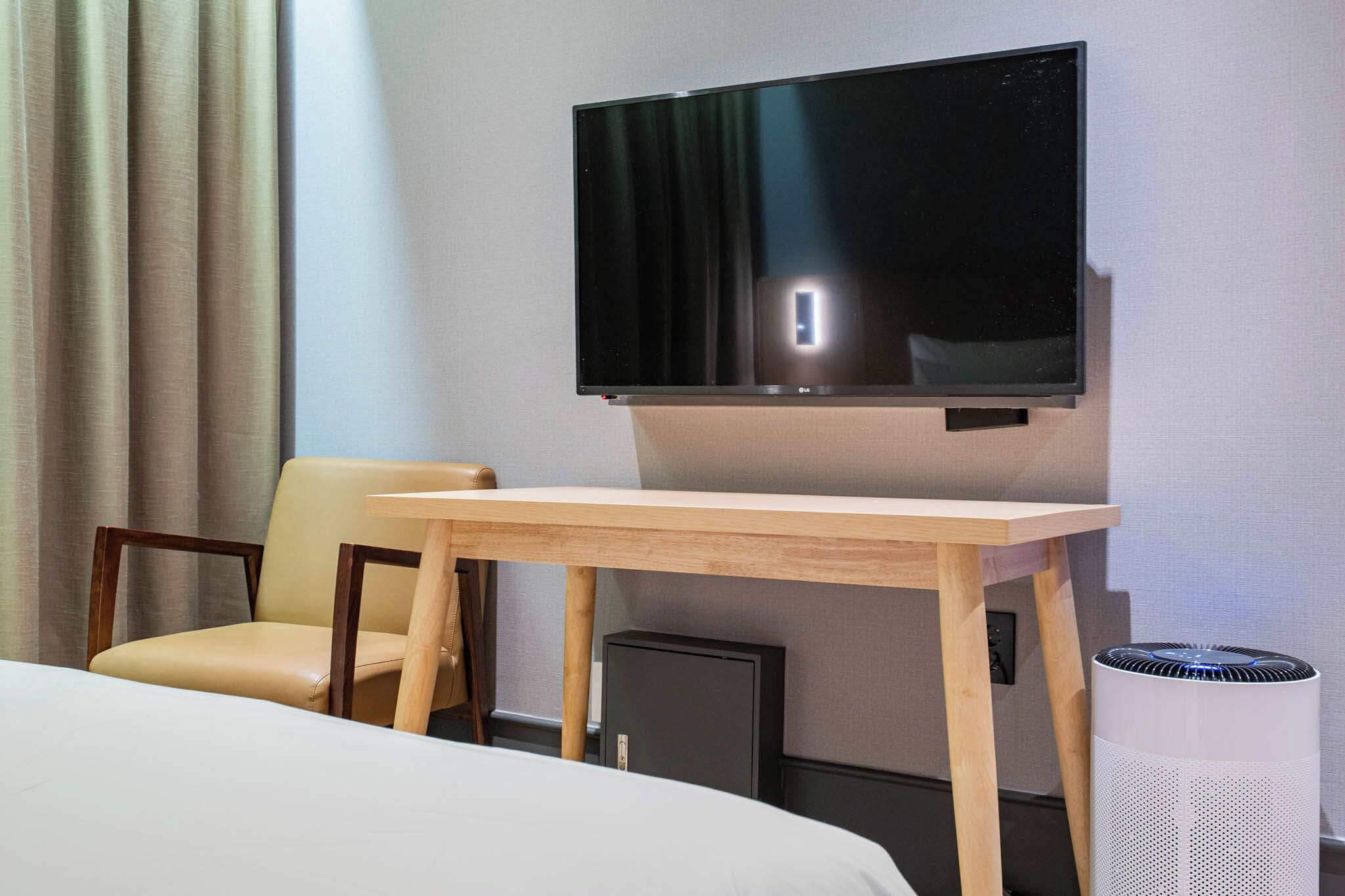 A hotel room with a bed, a wooden table, a TV mounted on the wall, a chair, and a waste bin.