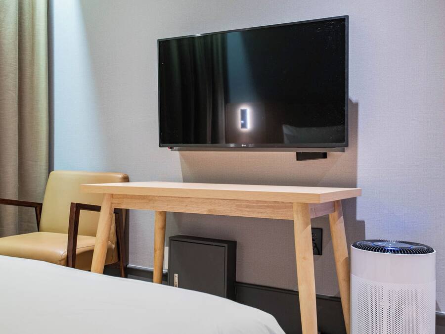 A hotel room with a bed, a wooden table, a TV mounted on the wall, a chair, and a waste bin.
