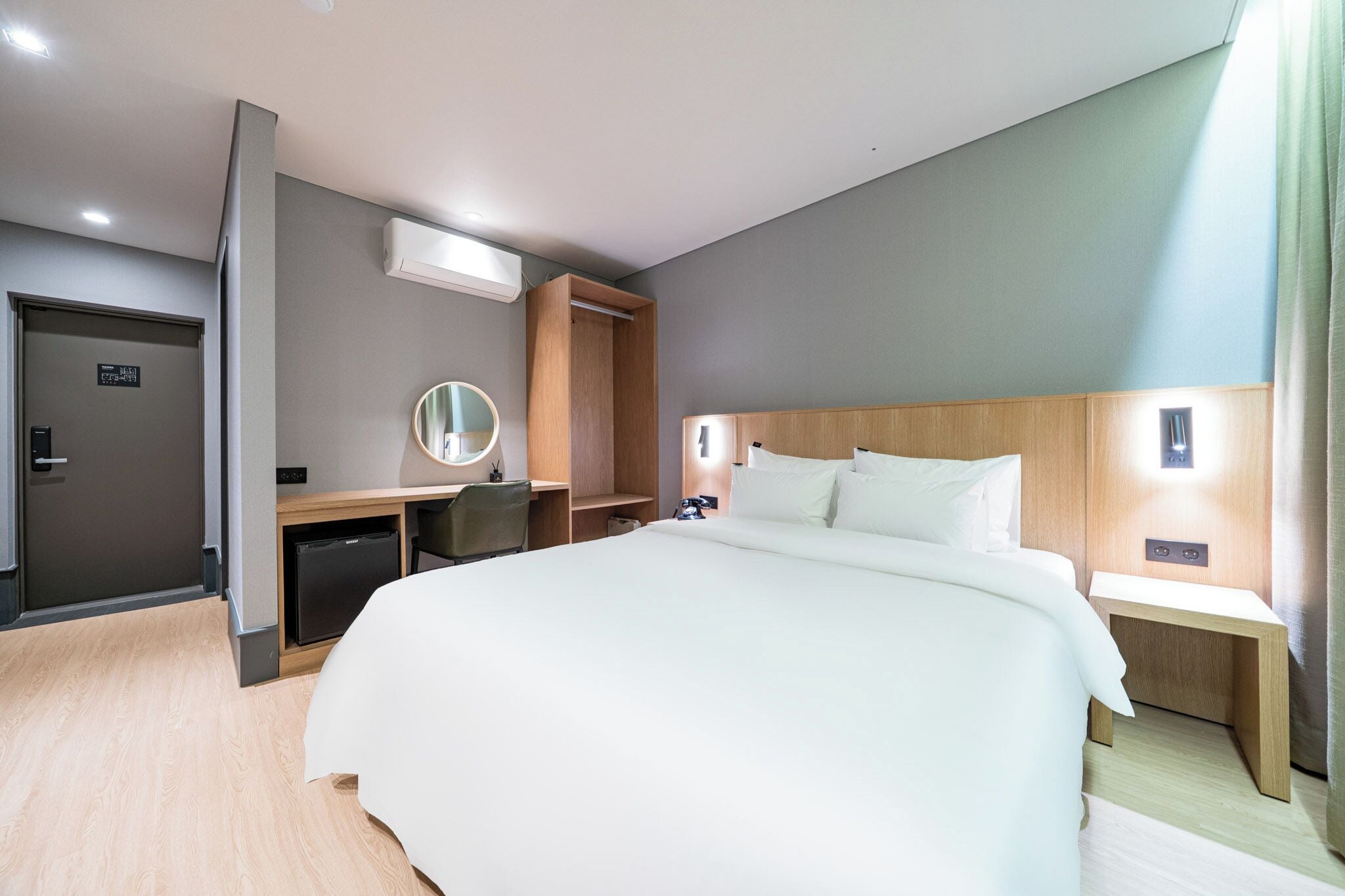 Ein modernes Hotelzimmer mit einem großen Bett, einem Schreibtisch mit Spiegel, einer kleinen Kühlschrank und einem Nachttisch.