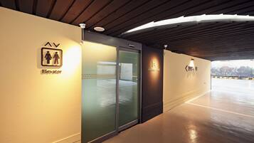 An elevator area with a sign indicating it is for men and women, a glass door entrance, and a view of a parking lot with a building in the background.