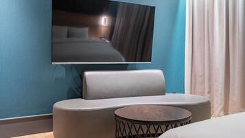 Una habitación de hotel moderna con una gran televisión de pantalla plana montada en la pared, un sofá gris, una mesita redonda de madera y una cama con ropa de cama blanca.